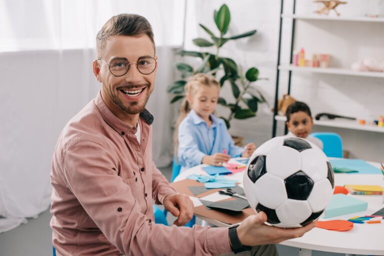 Smiling Teacher With Soccer Ball And Multicultural Classmates Behind In Classroom